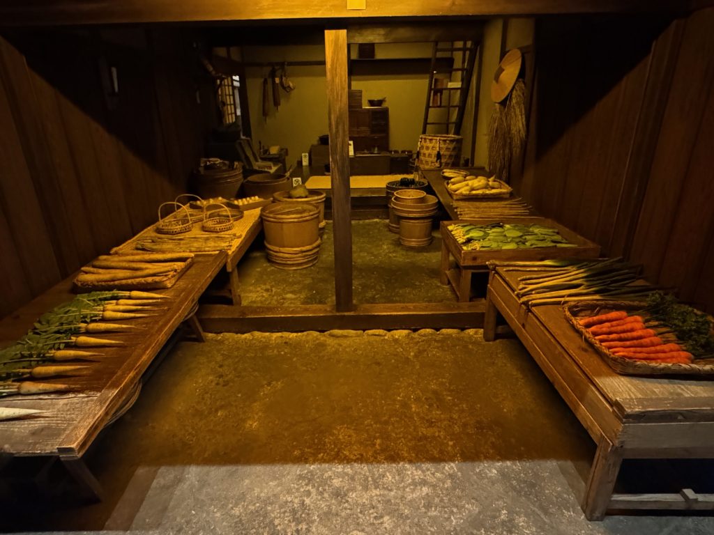 A photograph showing a recreation of a greengrocer's shop at the Fukagawa Edo Museum. Carrots, burdock, daikon, and pickled cabbage in barrels are shown.