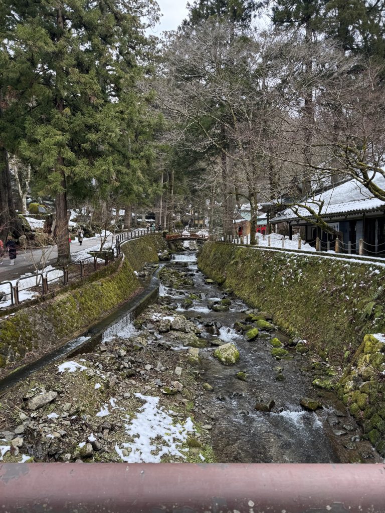 Looking up the rushing Eiheiji River, with stone wall-lined mossy banks bounded by trees and snow.