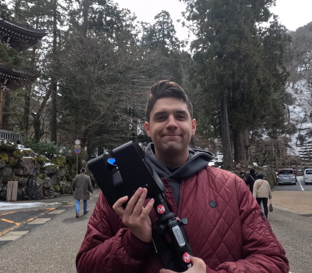 A student in a red jacket holding a Matterport Pro3 LiDAR scanner, which looks like a black shoe box, with trees in the background.