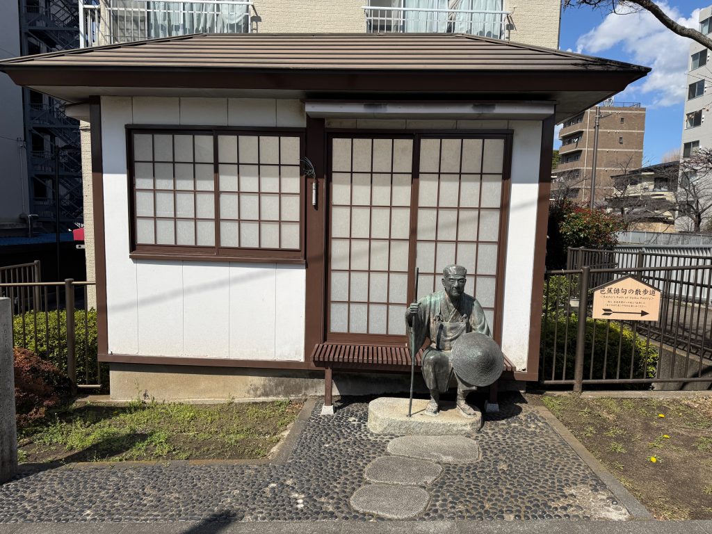A small Japanese-style house with a bronze statue of Bashō seated on a step in front of it.