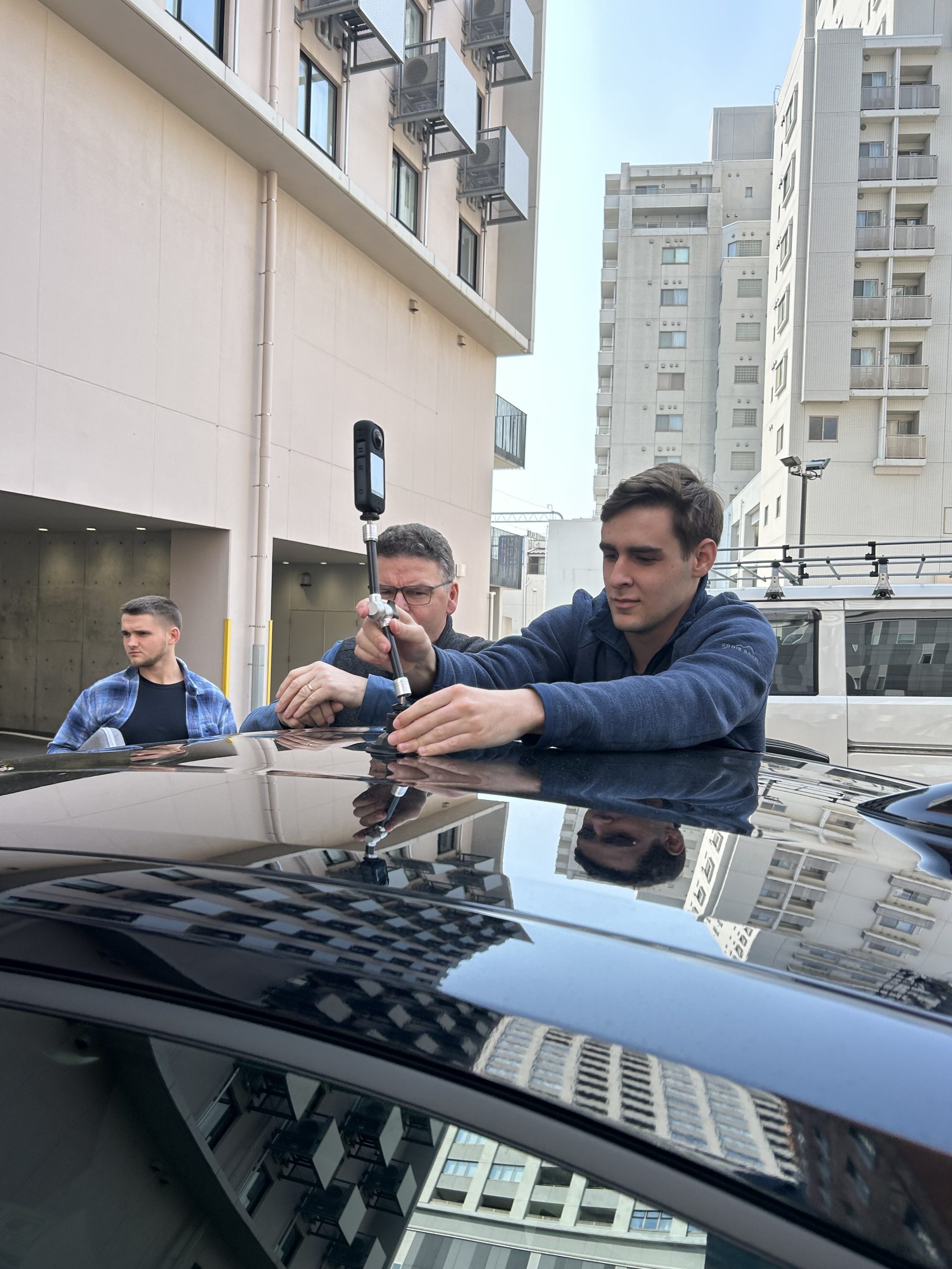 Two men mount a 360° camera on top of a car in a small parking lot in Fukui, Japan.