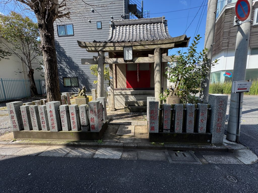 A small Shintō shrine, with traditional peaked and gabled roof, surrounded by dedication stones and guarded by stone foxes.